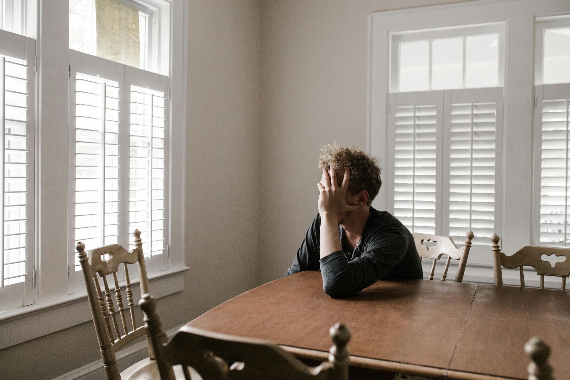 A man sits alone at a dining table with his head resting on his hand in an empty room